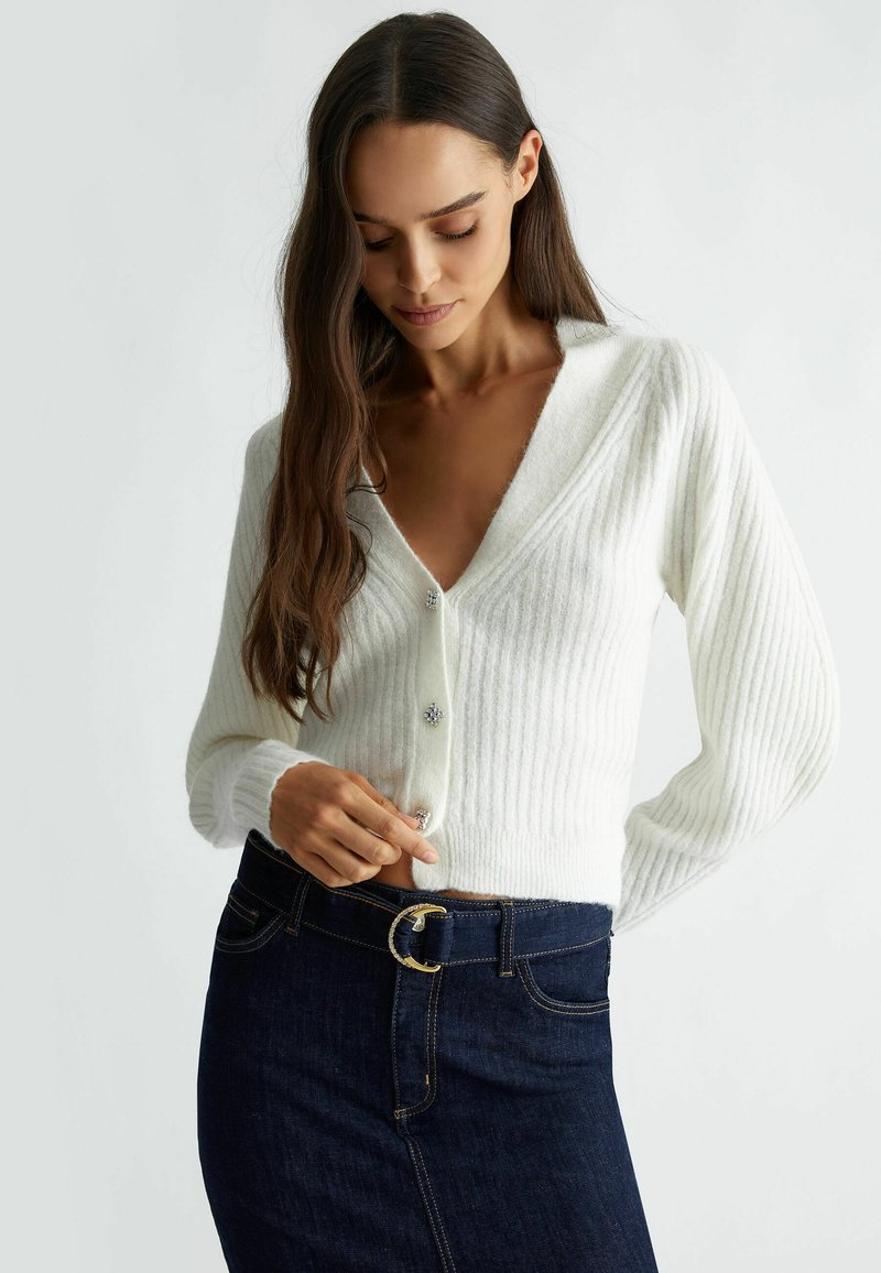 Cardigan bianco a coste con scollo a V e maniche lunghe, presenta chiusure decorative a bottoni. Abbinato a una gonna in denim scuro, con un elegante cinturino curvo.