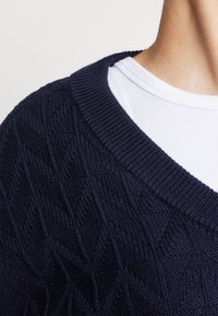 Gros plan sur une personne portant un pull en maille torsadée bleu marine par-dessus un t-shirt blanc côtelé à col rond.