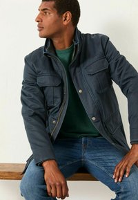 Marineblaue, Reißverschlussjacke mit vier Fronttaschen, hohem Kragen und Knopfakzenten, über einem grünen Pullover und blauen Jeans getragen.