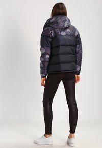 Kvinna klädd i en svart puffjacka med blommönster, svarta leggings och vita sneakers, stående vänd bort mot en enfärgad bakgrund.