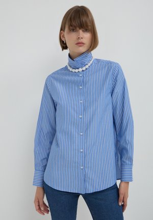 Woman wearing blue and white striped button-up shirt with high collar and large pearl necklace, paired with blue jeans against white background.