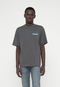 Graues kurzärmeliges Baumwoll-T-Shirt mit einem blauen "Paradise"-Grafikdesign im linken Brustbereich. Lockere Passform, Rundhalsausschnitt, glatte Textur.