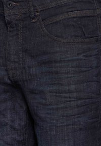 Tmavě modré džíny s mírně opotřebovanou texturou, s oranžovým prošitím a klasickým designem pěti kapsiček. Kovové zapínání na knoflík v pase.