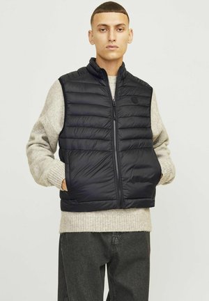 Gilet en duvet noir avec un col haut, fermeture éclair à l'avant et coutures horizontales. Porté par-dessus un pull en maille gris clair.