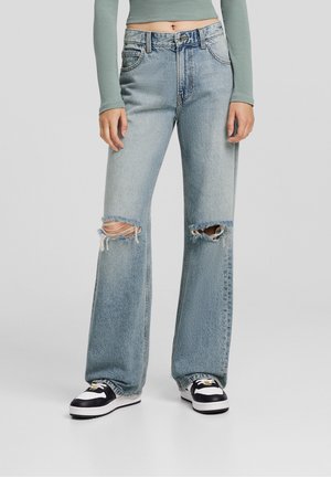 Light blue denim flared jeans with two large knee rips, five pockets, and a button closure. Paired with black and white sneakers.