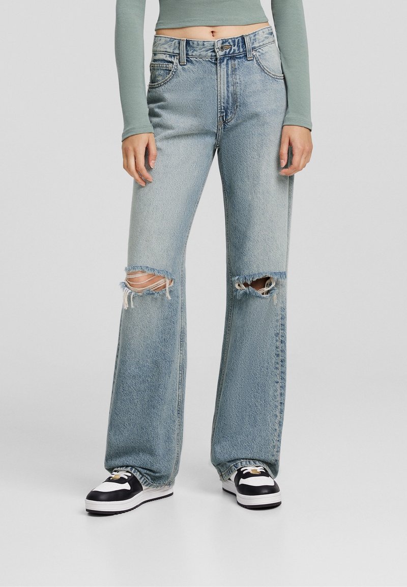 Light blue denim flared jeans with two large knee rips, five pockets, and a button closure. Paired with black and white sneakers.
