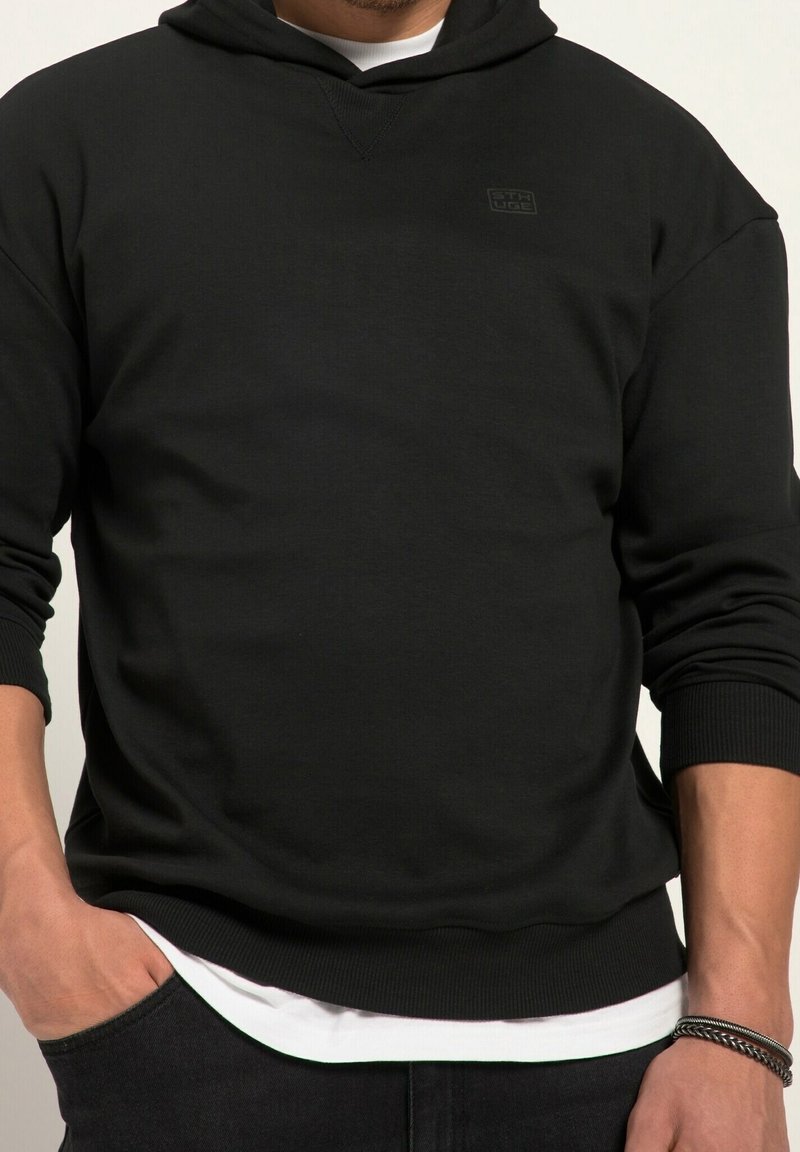 Homme portant un sweat à capuche noir avec un petit logo sur la poitrine, une chemise blanche en dessous, et un pantalon noir, main droite dans la poche, bracelet argenté au poignet.