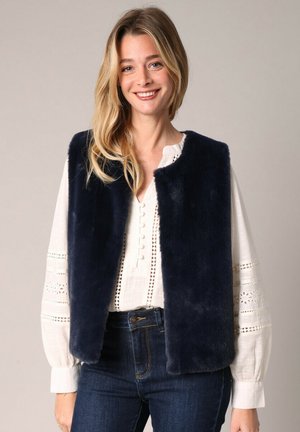 Veste sans manches en fausse fourrure bleu marine avec un col rond, des ouvertures à l'avant et une texture douce, portée par-dessus un chemisier blanc brodé et un jean sombre.
