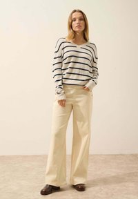 Sweter w paski w odcieniach kremowym i granatowym z dekoltem w kształcie litery V, zestawiony z kremowymi spodniami z szerokimi nogawkami z kordurowego materiału, które mają boczne guziki i fakturowaną powierzchnię.