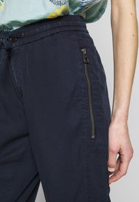 Marineblauwe broek met een elastische tailleband, trekkoord en zijzak met rits. Het materiaal lijkt lichtgewicht met een gladde textuur.