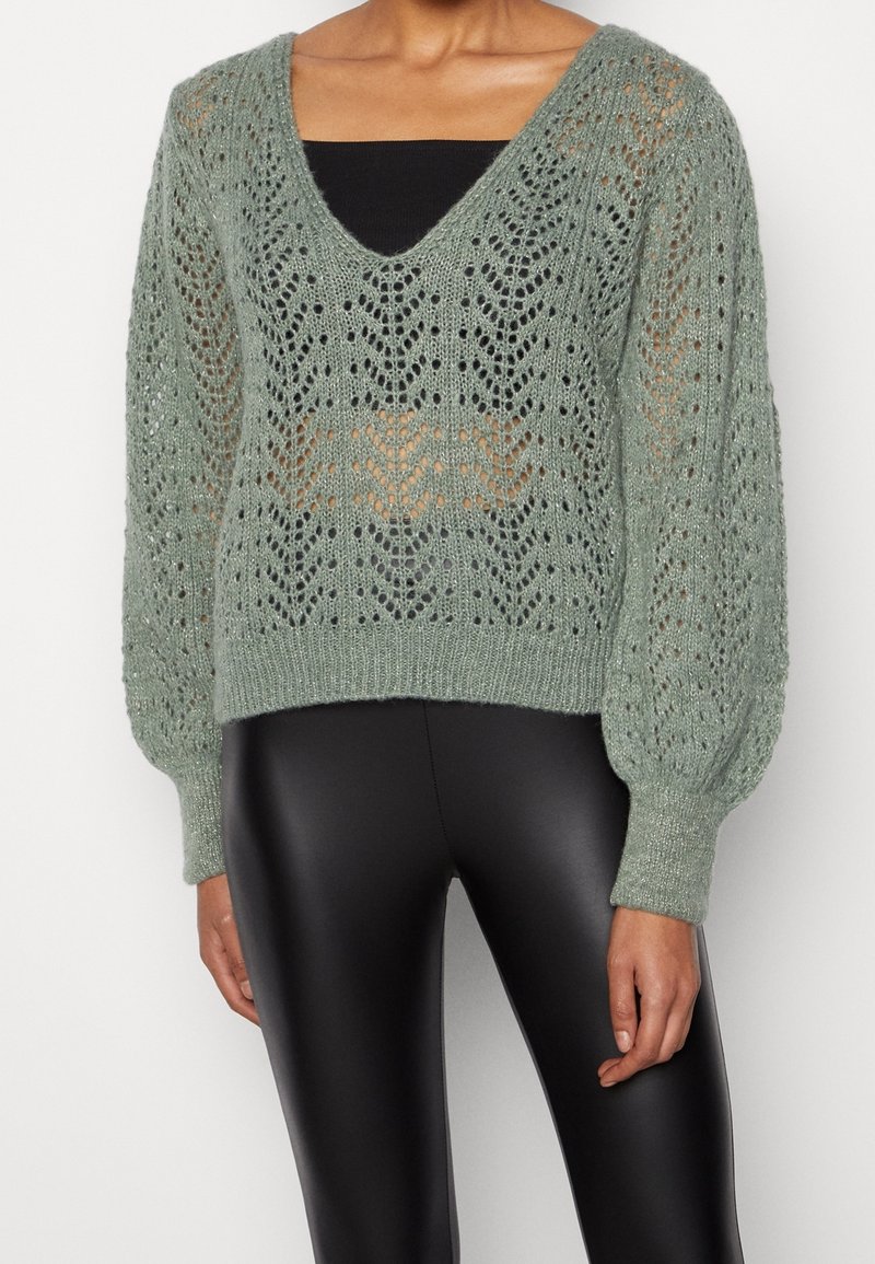 Femme portant un pull en maille vert clair à col en V avec un motif en dentelle florale et des manches bouffantes, par-dessus un haut noir et un pantalon en similicuir noir.