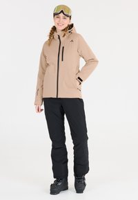Beige vattentät jacka med dragkedja fram, svarta detaljer och huva; i kombination med svarta skidbyxor och svarta skidstövlar; goggles på huvudet.