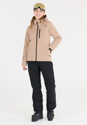 Beige waterdichte jas met een rits aan de voorkant, zwarte accenten en capuchon; gecombineerd met zwarte skibroek en zwarte skischoenen; skibril op het hoofd.