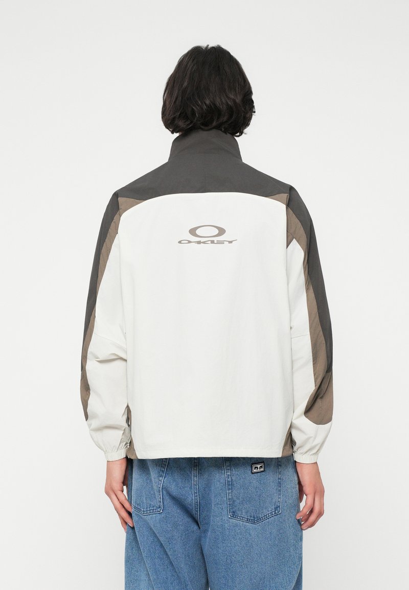 Veste coupe-vent blanche et gris foncé avec des poignets élastiques, arborant le logo "Oakley" sur le haut du dos et des panneaux d'accent marron.