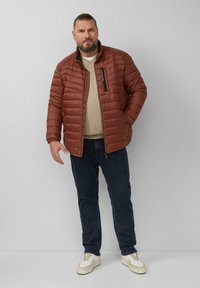 Bruine gewatteerde jas met een hoge kraag, ritssluiting en een borstzak. Draagt over een beige trui met donkerblauwe jeans en witte sneakers.