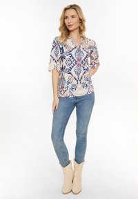 Blusa leggera e colorata con motivo floreale e geometrico, maniche corte, scollatura a V, indossata con jeans attillati e stivali da cowboy beige.