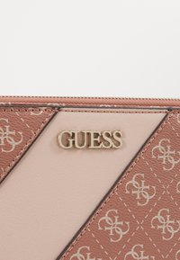 Brązowy i jasnopudrowy portfel o geometrycznym wzorze, z złotym logo "GUESS" i teksturowanym wykończeniem. Zamek błyskawiczny na górze.