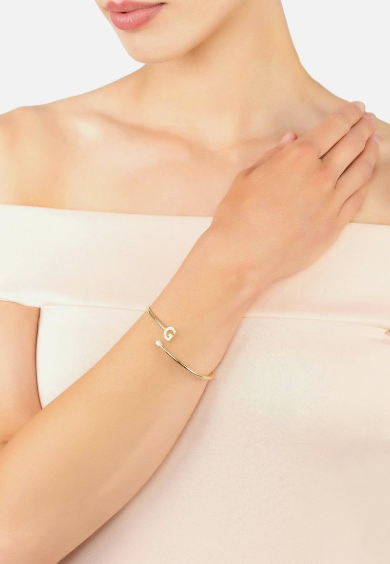 Gold bracelet with a circular design, featuring a letter "G" and a small diamond accent. Worn on the wrist against a skin tone background.