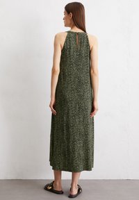Maxikleid in Dunkelgrün mit verstreutem weißen Muster, ärmelloses Design, Detail mit Schlitz im Rücken und fließendem Stoff. Flache Sandalen getragen.