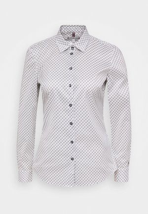 Witte blouse met lange mouwen en knopen, voorzien van een zwart rond bloemmotief en een spitse kraag, afgebeeld tegen een effen achtergrond.