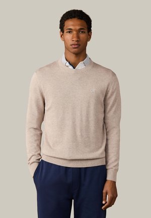 Jonge man draagt een beige trui met ronde hals over een wit overhemd en donkerblauwe broek, staand tegen een neutrale achtergrond met handen in de zakken.