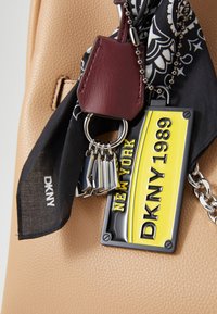 Hnědá kožená kabelka se stříbrnými doplňky, černý šátek s bílými vzory a žlutý a černý klíčový řetízek DKNY s designem z roku 1989.