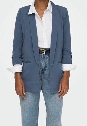 Person trägt einen blauen Blazer über einem weißen Hemd mit Knöpfen, das in eine hoch taillierte hellblaue Jeans mit schwarzem Gürtel gesteckt ist.
