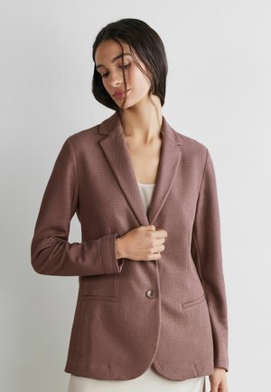 Frau mit dunklem Haar, die einen mauvefarbenen Blazer trägt, am Bund geknöpft, stehend vor einer schlichten weißen Wand.