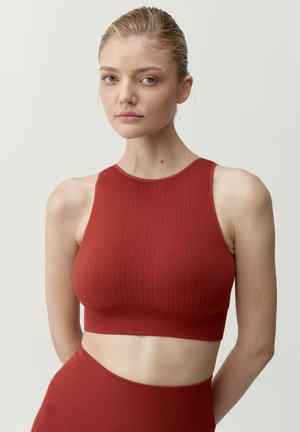Młoda kobieta o blond włosach w czerwonym, teksturowanym, bezrękawnikowym topie odsłaniającym brzuch oraz pasujących, wysokich spodenkach na tle jednolitego, jasnego tła.