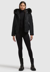 Black puffer jacket with a fur-trimmed hood, zip closure, and two side pockets. Worn over a black turtleneck and black leggings.