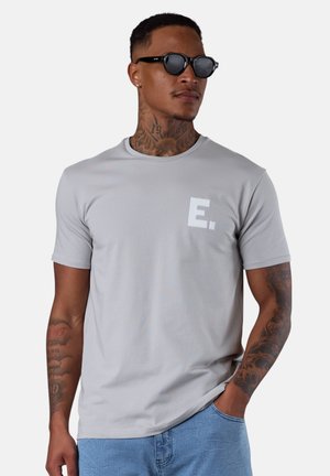 Light gray crew-neck t-shirt made of smooth fabric, featuring a textured white “E.” logo on the chest. Model also wears black sunglasses.