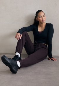 Schwarzes Langarmshirt und maroonfarbene Jogginghosen. Das Outfit hat eine körpernahe Passform und wird mit schwarzen Sneakers und weißen Socken kombiniert, während die Person an einer Wand sitzt.