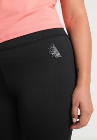 Sorte leggings med et tætsiddende design, der har et reflekterende sølvlogo, parret med en lys pink top. Den glatte stofstruktur er synlig.