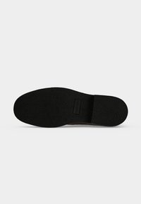 Semelle en caoutchouc noire texturée d’une chaussure avec un talon légèrement surélevé et un logo rectangulaire embossé près de la zone de la voûte plantaire.
