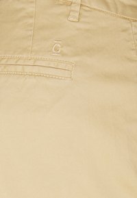 Beige bomullbyxor med en slät textur, med en ficka med en sömnadsdetalj och en diskret logotyp på tyget.