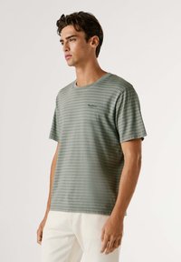 Jeune homme portant un t-shirt à manches courtes vert à rayures et un pantalon blanc, debout et regardant vers la gauche sur un fond uni.