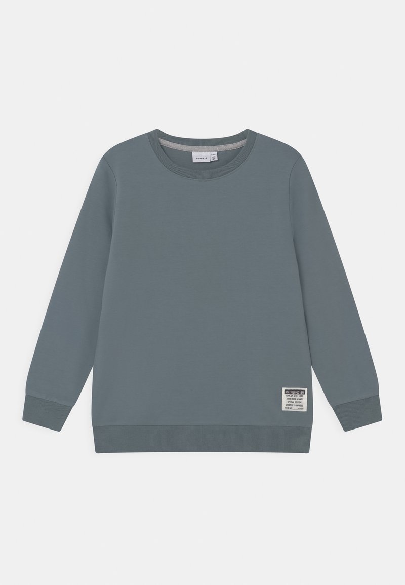 Sweatshirt gris à manches longues, poignets côtelés et encolure ronde. Présente une étiquette sur l'ourlet avec des instructions d'entretien et le logo de la marque. Texture lisse.