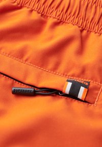 Tejido elástico naranja con cintura fruncida, que presenta un cordón negro. Incluye una etiqueta negra y naranja con el texto "SUPERDRY".