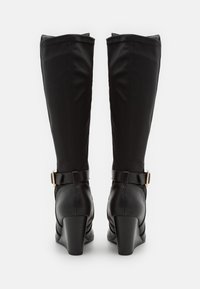 Une paire de bottes noires à talons compensés, montant jusqu'aux genoux, avec des sangles de boucle à l'arrière et des tiges à texture lisse sur fond blanc.