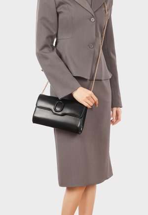 Donna in completo color taupe che tiene una borsa a tracolla in pelle nera con catenella dorata su uno sfondo uniforme.