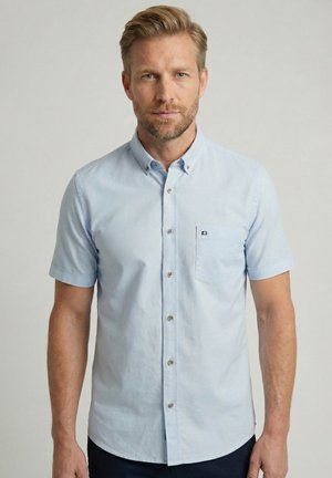 Man met kort blond haar en baard, gekleed in een lichtblauw kort mouw overhemd, staand tegen een effen achtergrond.