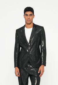 Zwarte paillettenblazer met een getailleerde pasvorm, enkele knoopsluiting en twee voorkruizen. Gecombineerd met bijpassende paillettenbroek.