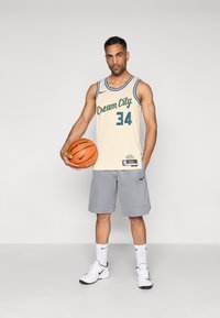 Basketbalový dres Cream City v svetlej béžovej farbe s zeleným nápisom, číslo 34, v kombinácii so sivými šortkami Nike a bielymi teniskami, držiaci basketbal.