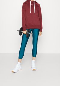 Felpa bordeaux con cappuccio e coulisse, leggings turchesi e sneakers bianche. Modello tiene in mano un manubrio nero, su uno sfondo grigio chiaro.