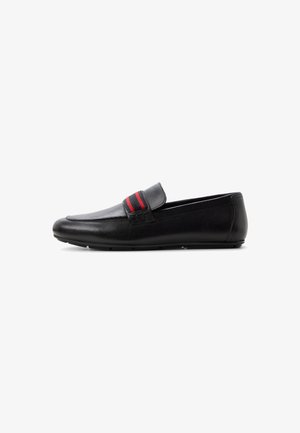 Mocassin en cuir noir à semelle plate, avec une bande rayée rouge et noire sur le dessus, vu de côté sur un fond blanc.