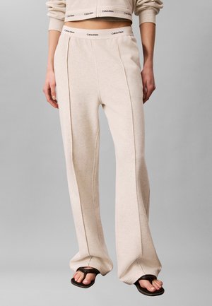 Béžové tepláky Calvin Klein s detailmi švov, nosené s originálnou krátkou mikinou a čiernymi sandálmi so štvorcovou špičkou na sivom pozadí.