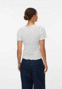Kvinna med hår i en knut, klädd i en vit texturerad kortärmad topp och mörkblå byxor, sedd bakifrån mot en vit bakgrund.