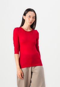 Red long-sleeve shirt made of smooth fabric, featuring a round neckline and fitted design. Paired with loose beige pants.