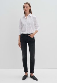 Chemise blanche à boutons avec des manches longues, associée à un jean noir slim. Le mannequin porte des chaussures noires à enfiler et se tient devant un fond clair.