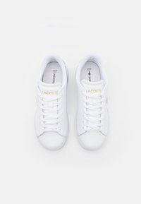 Paire de baskets blanches Lacoste avec logo doré sur la languette, lacets blancs et semelles intérieures brandées sur fond blanc uni.